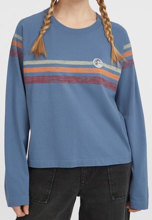 Personne portant une chemise bleue à manches longues avec des rayures horizontales crème, orange et rouge, associée à un pantalon noir, cheveux coiffés en deux tresses.