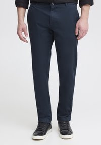 Muž v tmavě modrých slim-fit kalhotech a černých teniskách s bílou podrážkou, stojí na pozadí jednobarevné světlé plochy.