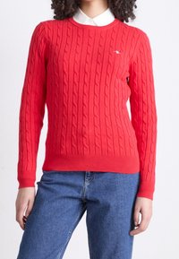 Femme portant un pull rouge vif en maille torsadée par-dessus une chemise blanche à col et un jean en denim bleu, sur un fond uni.