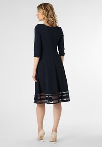 Dunkelblaues Kleid mit Dreiviertelärmeln, einem figurbetonten Oberteil und einem ausgestellten Rock, der am Saum ein transparentes Paneel mit horizontalen Streifen aufweist.