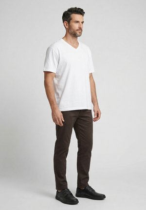 Man staat, draagt een wit T-shirt met V-hals, donkerbruine broek en zwarte veterschoenen tegen een effen lichtgrijze achtergrond.