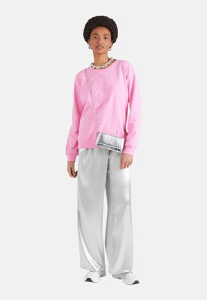 Vrouw staat en draagt een roze sweatshirt, zilveren wijde broek, witte sneakers, een zilveren ketting, een zilveren crossbody tas en oorbellen.