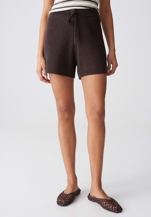 Frau trägt dunkelbraune gestrickte Shorts mit Kordelzug und passenden braunen gewebten Slippern, steht vor einem schlichten hellen Hintergrund.