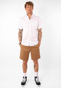 Chemise blanche à manches courtes avec de petits pois bleus et rouges ; poche poitrine. Shorts marron ; baskets noires avec accents blancs ; chaussettes blanches.