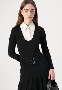 Fekete, hosszú ujjú bodi, mély V-kivágással, amely alatt fehér galléros ing van. Pliszézett fekete szoknya és fekete öv, téglalap alakú csattal.