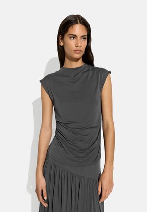 Młoda kobieta z długimi ciemnymi włosami, ubrana w ciemnoszary bezrękawnik z marszczeniami i plisowaną spódnicę, stojąca na tle jednolitego jasnego tła.