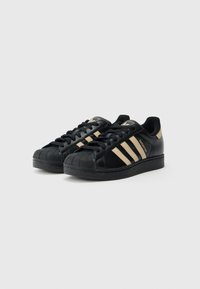 Černé tenisky Adidas Superstar s béžovými pruhy a špičatým designem, položeny vedle sebe na jednobarevném bílém pozadí.