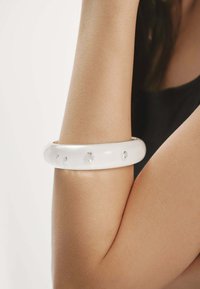Brazalete bangle blanco hecho de un material brillante, que presenta piedras transparentes incrustadas en la parte frontal, llevado en el brazo de una persona.