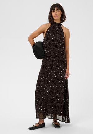 Frau trägt ein ärmelloses schwarzes Maxikleid mit weißen Punkten, hält eine schwarze Clutch und trägt schwarze Ballerinas mit Knöchelriemen.
