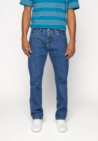 Vaqueros de mezclilla azul con corte recto, que cuentan con cinco bolsillos, bragueta con botones y un sutil desgaste. Combinados con una camiseta de rayas azules y blancas.