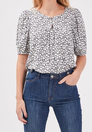 Vrouw met een witte en zwarte bloemenblouse met korte mouwen, gestopt in een high-waisted blauwe jeans, hand in de zak, tegen een effen achtergrond.