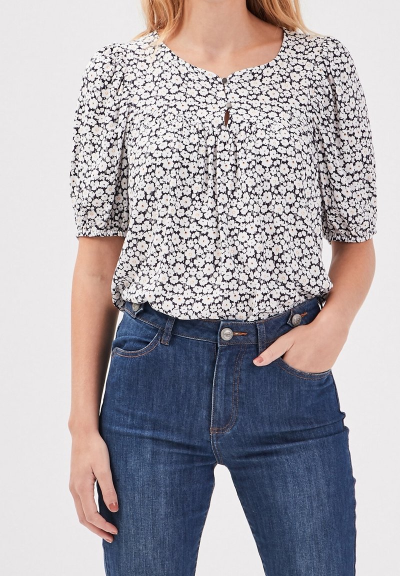 Femme portant un chemisier à manches courtes blanc et noir à motif floral, rentré dans un jean bleu taille haute, main dans la poche, sur un fond uni.