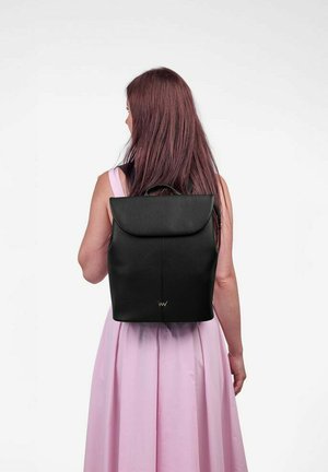 Černý kožený batoh s kulatým víkem, minimalistický design a jemné detaily prošívání. Nesený na zádech přes růžové šaty.