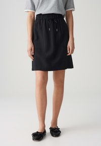 Frau trägt einen schwarzen knielangen Rock mit Kordelzugbund, ein graues Kurzarmshirt und schwarze Ballerinas mit Schleifen, stehend vor einem schlichten Hintergrund.