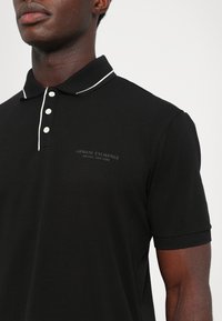 Svart polo-skjorte med klassisk krage, tre-knapps placket og hvite detaljer. Har et brodert merke-logo på brystet. Myk stoff.