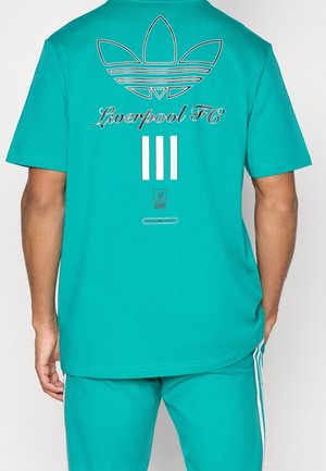 Turkusowa koszulka z logo Adidas, tekstem "Liverpool FC" i trzema białymi paskami na plecach, noszona z dopasowanymi turkusowymi spodniami.