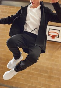 Schwarze Sportjacke über einem weißen T-Shirt, kombiniert mit schwarzen Jogginghosen und weißen Turnschuhen, in einer Halle für Basketball.