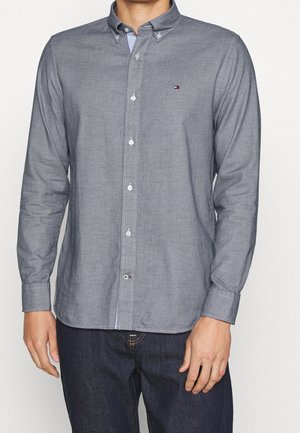 Homme portant une chemise grise à manches longues boutonnée avec un petit logo sur la poitrine et un jean bleu foncé, debout devant un fond uni.