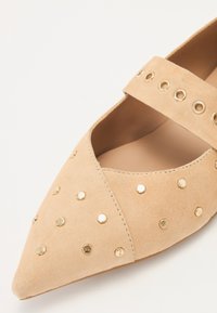 Pantofii plata din suede bej, cu vârful ascuțit, dotati cu ținte decorative aurii și o curea înclinate pe glezna piciorului. Căptușeală interioară netedă.