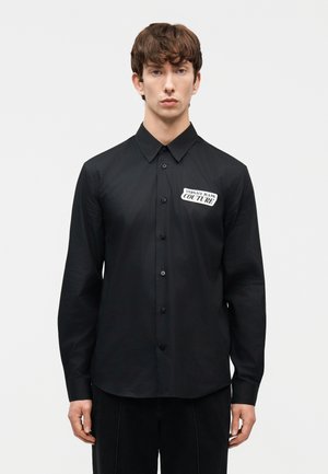 Mees, kes kannab musta nööbitavat särki, mille vasakul rinnal on "Versace Jeans Couture" logo, seisab lihtsa heleda tausta ees.