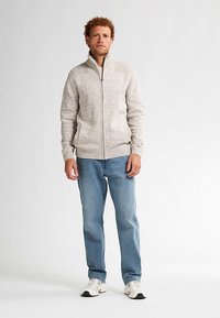 Světlounce šedý svetr na zip s žebrovanými detaily, pletenou texturou a dvěma bočními kapsami, doplněný modrými džíny a bílými teniskami.