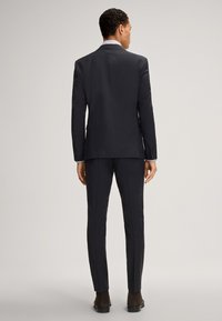 Marineblaues Anzugensemble mit einem taillierten Sakko, das über einen mittigen Schlitz verfügt, und passenden Hosen. Der Stoff wirkt glatt; das Sakko hat eine strukturierte Silhouette und Knöpfe.