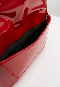 Sac à main rouge brillant avec un rabat ouvert, dévoilant une doublure intérieure noire lisse et une attache en chaîne dorée à l'intérieur.