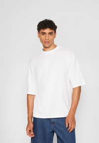 Tricou alb din bumbac cu mâneci scurte, guler rotund, croială lejeră. Asortat cu pantaloni scurți din denim albastru. Textură netedă, design minimalist.
