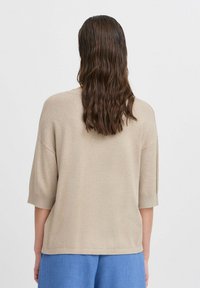 Beige pletený svetr s krátkými rukávy a uvolněným střihem, má kulatý výstřih a postranní rozparky, v kombinaci se světle modrými kalhotami.