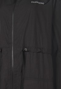 Chaqueta ligera negra con cremallera frontal, cintura ajustable con cordón y bolsillos laterales. Presenta una textura suave y un sutil logo en el cuello.