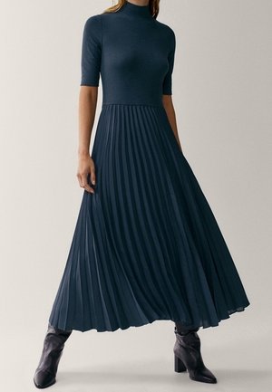 Femme portant une robe midi plissée bleu marine à manches mi-longues et des bottines à talons noires, debout devant un fond uni.