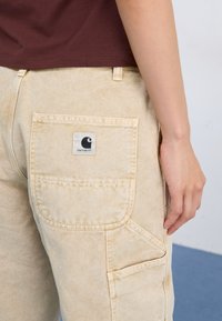Beige arbetsbyxor med en stor, fyrkantig bakficka som har en svart Carhartt-logotyp. Texturen är mjuk, med synliga sömmar och förvaringsdetaljer.