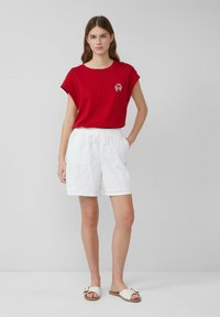 Vrouw in een rode mouwloze top met een krab embleem, witte oogjes shorts en witte instappers, staande tegen een lichtgrijze achtergrond.