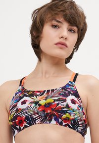 Soutien-gorge de sport floral avec une base noire, ornée de motifs tropicaux vibrants en rouge, jaune et vert ; bretelles réglables et détails de logo.