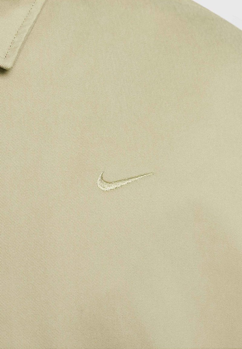 Camisa polo de tejido en color beige claro con un acabado texturizado. Presenta un pequeño logo bordado de Nike en forma de swoosh en un sutil tono dorado.