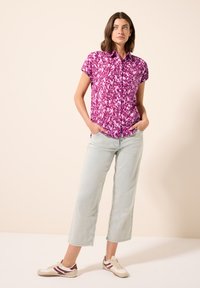Fuchsia kortebloes met knopen en een geometrisch patroon, gecombineerd met lichtgewassen wijde jeans en beige sneakers met bordeauxrode accenten.