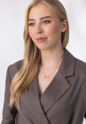 Blonde Frau mit langen Haaren, die einen braunen Blazer und eine goldene Halskette mit Buchstaben- und Herzanhängern trägt und leicht zur Seite schaut.