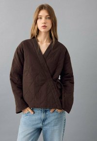 Braune, gesteppte Wickeljacke mit V-Ausschnitt, langen Ärmeln und seitlicher Bindung. Gepaart mit hellblauen Denim-Jeans. Strukturierter Muster auf dem Stoff sichtbar.