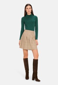 Grünes Langarm-Turtleneck-Oberteil mit subtilem Muster, kombiniert mit einem beigen karierten Faltenrock und knielangen braunen Suede-Stiefeln mit Absatz.