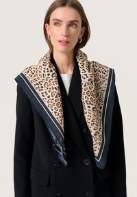 Løveplet-scarf i creme og brun, indrammet af marineblå kanter. Båret over en sort dobbeltbrystet frakke. Letvægts, tekstureret stof.