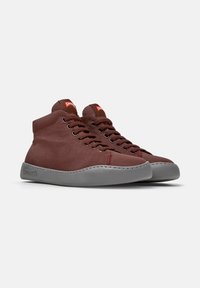 Chaussures montantes marron avec semelles grises, dotées de lacets et du logo Camper sur des étiquettes rouges. Positionnées sur un fond blanc uni.