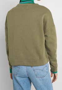 Sudadera verde oliva con un corte relajado, detalles de puños acanalados y un escote redondeado, combinada con jeans azul claro. Textura suave.