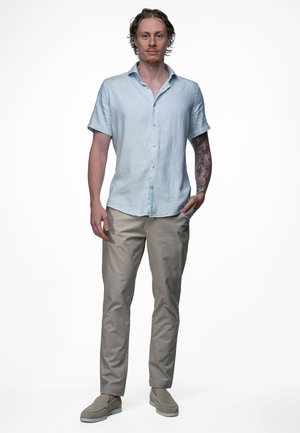 Man met een getatoeëerde rechterarm, die een lichtblauw shirt met korte mouwen draagt, beige broek en beige instapschoenen, staand met één hand in zijn zak.