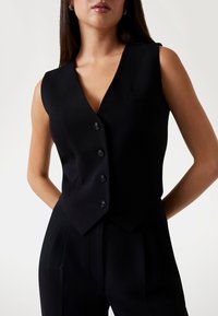 Black sleeveless vest with a deep V-neck, five button closure, and a chest pocket. Smooth fabric with a tailored fit.