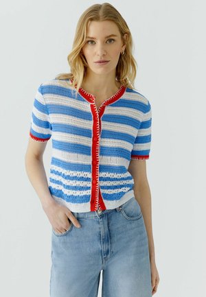 Femme portant un cardigan tricoté rayé bleu et blanc avec une bordure rouge et un jean taille haute bleu clair, debout devant un fond uni.