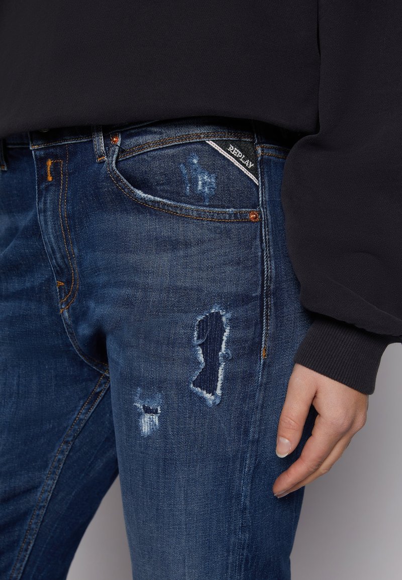 Jean en denim bleu foncé avec des détails usés, comprenant des patchs effilochés et un design classique à cinq poches. Étiquette de logo visible près de la poche avant.
