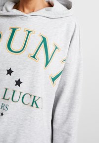 Grå hoodie med en stor grön och gul grafik som visar ordet "LUCKY" och svarta stjärnor. Mjukt tyg med avslappnad passform och huva.