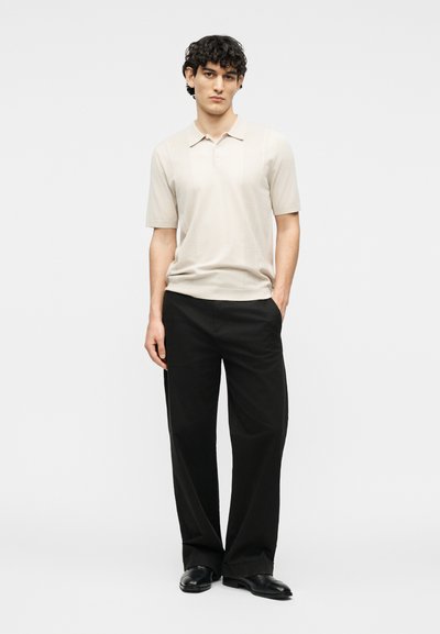 Νέος άνδρας με σγουρά σκούρα μαλλιά, που φοράει μπεζ πόλο μπλουζάκι με κοντά μανίκια, μαύρο παντελόνι με φαρδιά πόδια και μαύρα παπούτσια, στέκεται με το χέρι στην τσέπη.