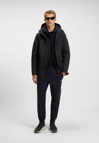 Black padded jacket with a hood, worn over a black sweatshirt and black joggers. Black sneakers with a light sole. Man wearing sunglasses.