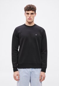 Čierna mikina s okrúhlym výstrihom, dlhými rukávmi, rebrovanými manžetami a lemom, s malým vyšívaným logom na hrudi, nosená s svetlomodrými džínsami.